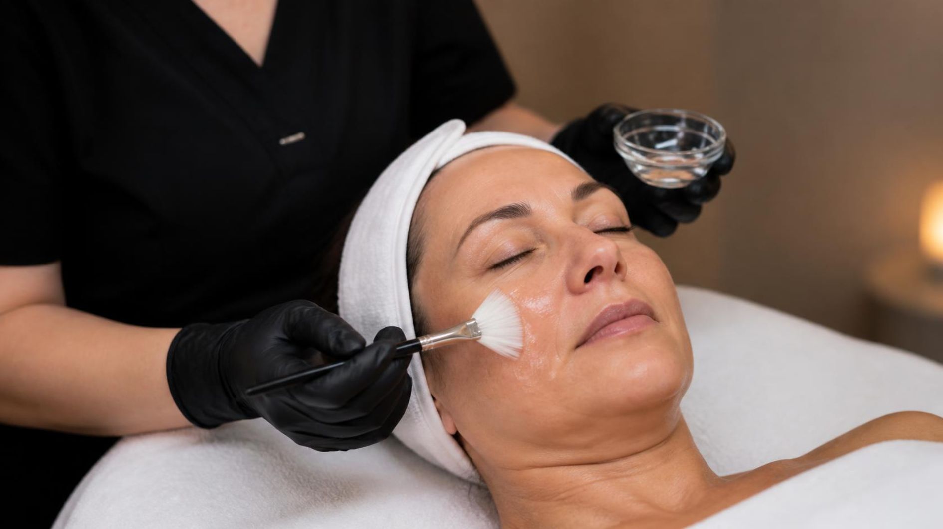 Esthetician in black scrubs applying a chemical peel to a relaxed client